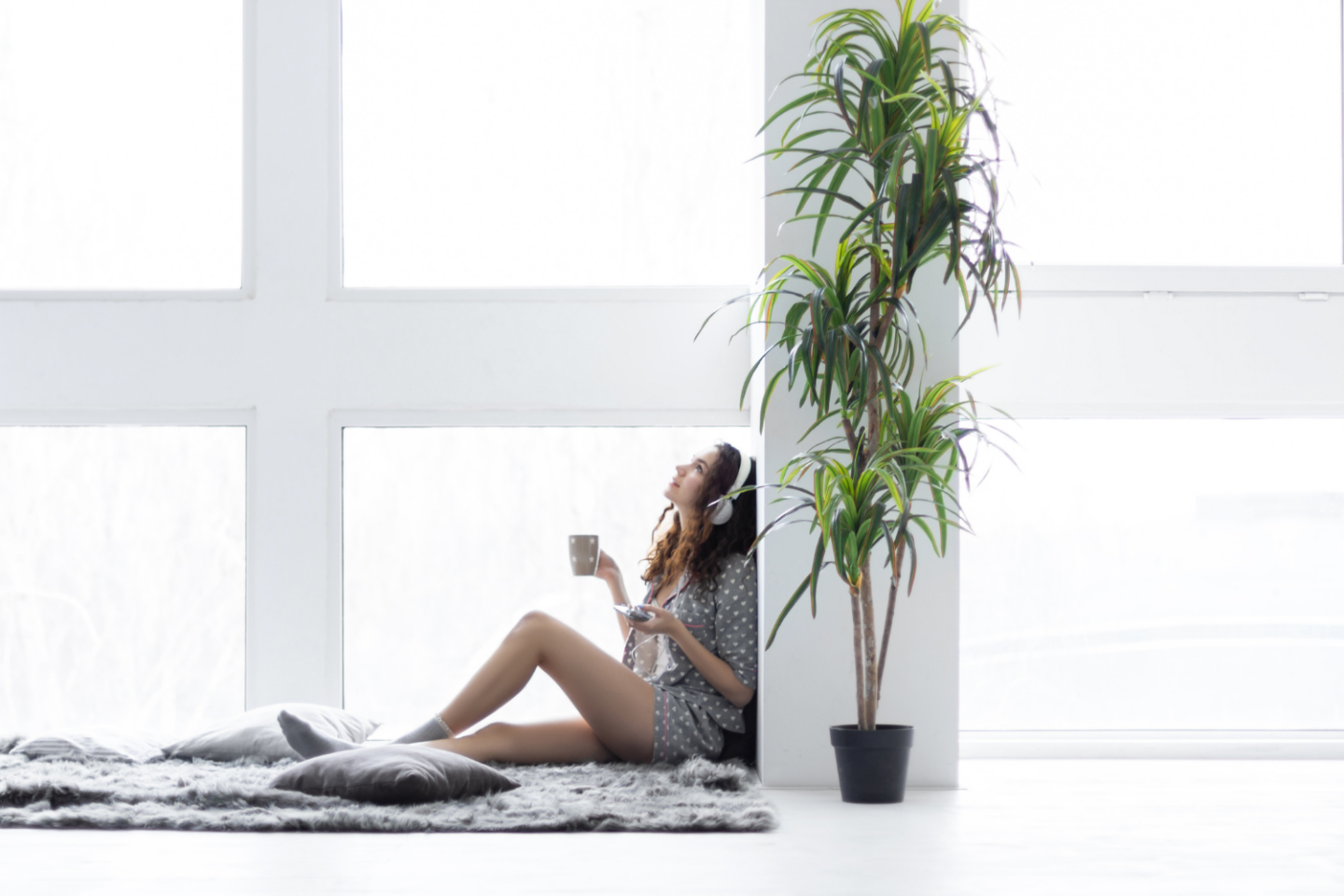 Woman sitting on the floor with her back to a column listening to music and contemplating productive morning routinesLocation is indoors in a white room with a large green plant.