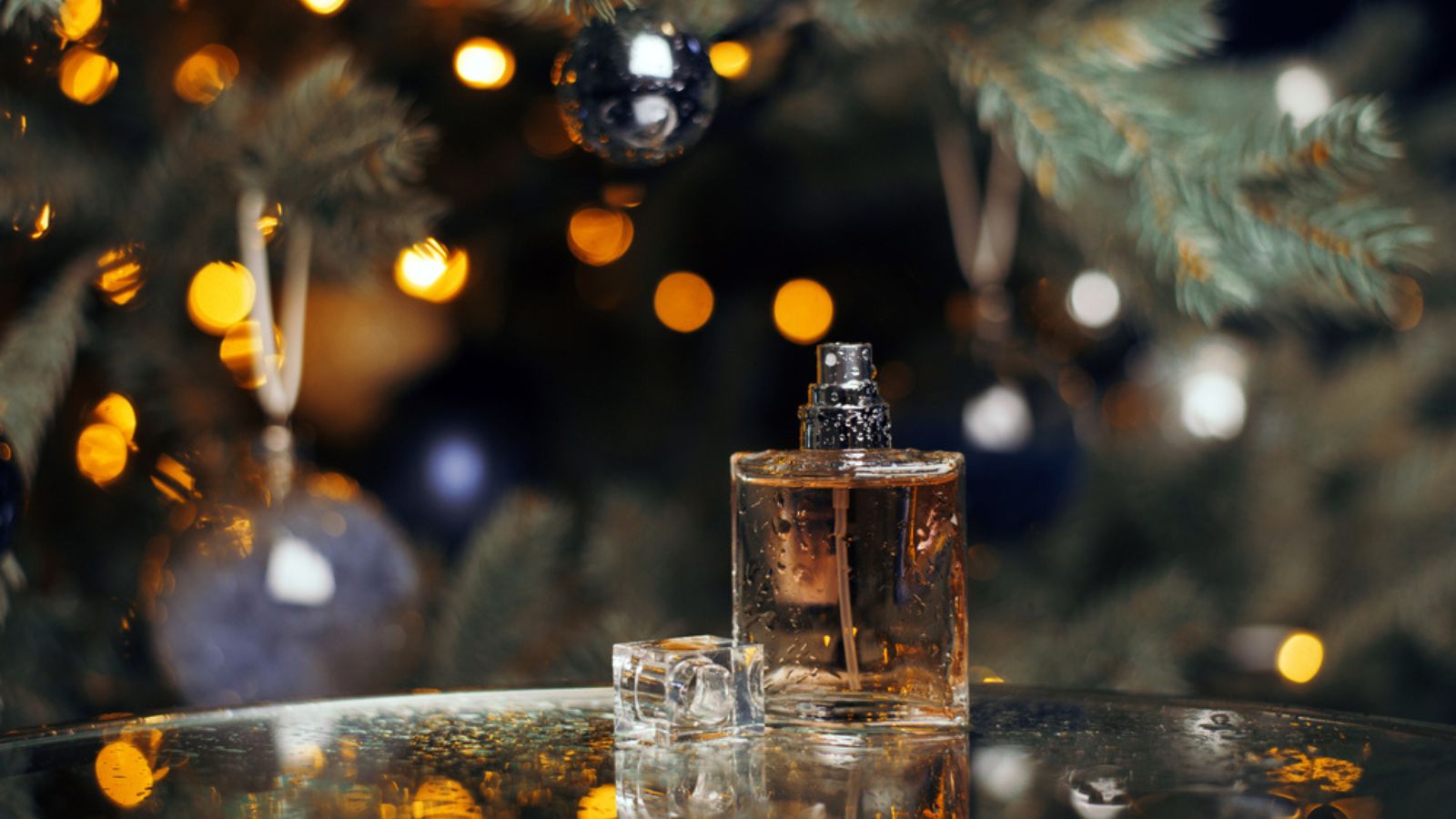 Beautiful composition of a bottle of women's perfume with drops of water on a glass table against the background of a Christmas tree