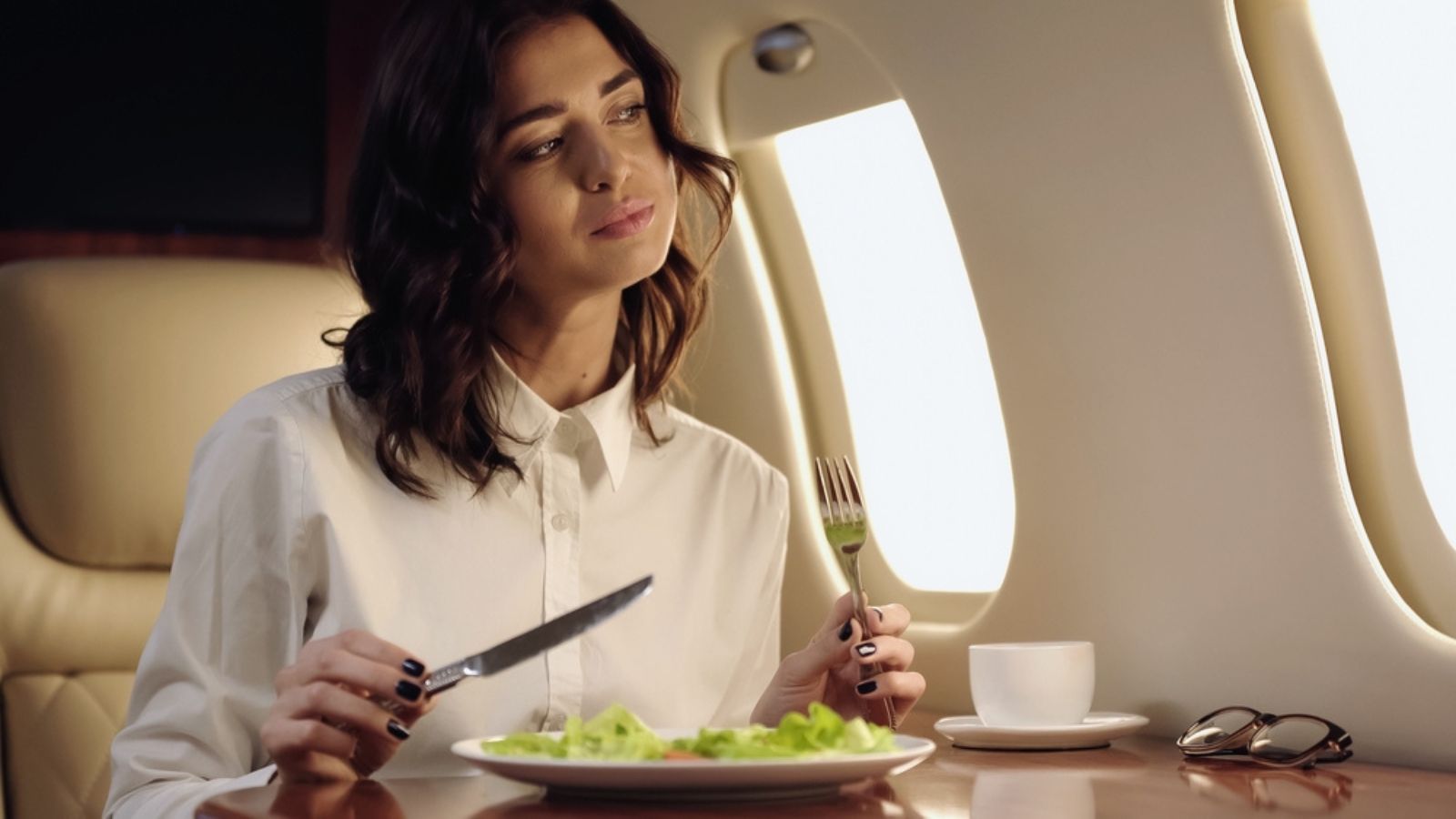 Pleased businesswoman holding cutlery near fresh salad and cup in private jet