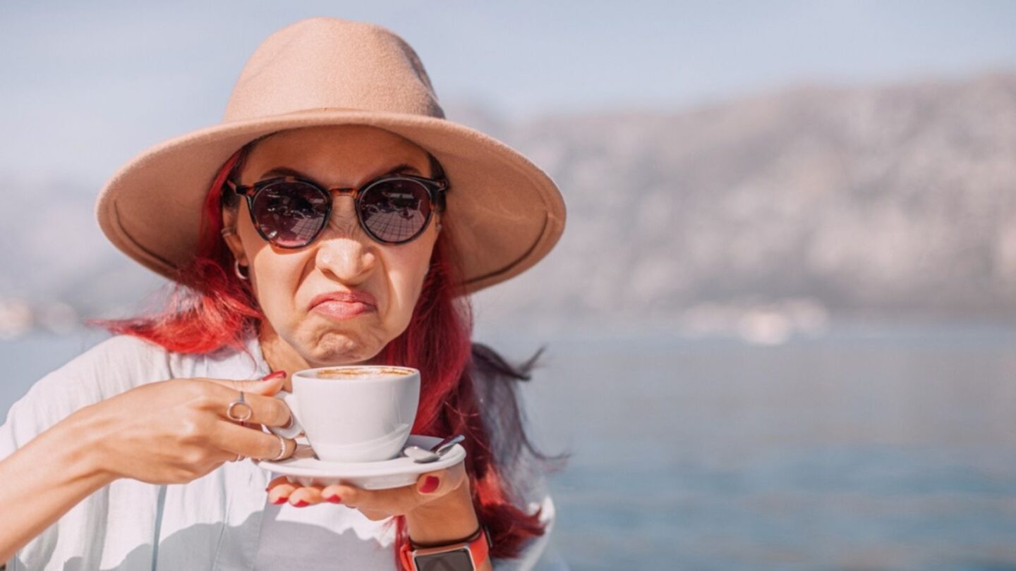 Redhead tourist with hat and sunglasses has a bad coffee experience at a restaurant by the sea